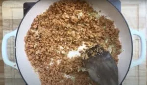 Pan with browned textured vegetable protein and onions cooking, stirred with a spatula on a wooden countertop.